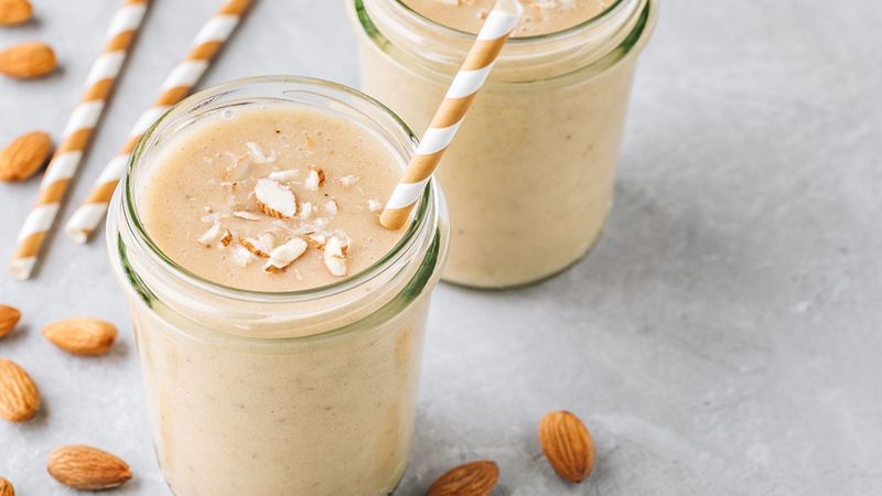Two mugs of dry fruit milkshakes with straws in them alongwith some almonds aside