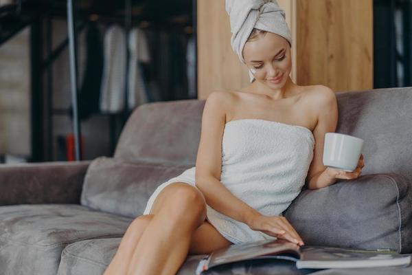 Indoor shot of relaxed beautiful woman sits relaxed on sofa, wears bath  towel around body after taking shower, holds cup of tea, reads magazine,  spends free time at home. Wellness, hygiene concept